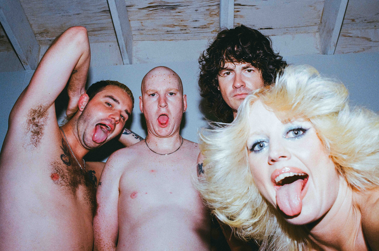 Australian punk rock band Amyl and The Sniffers posing together indoors with expressive faces and tongues out in a close-up portrait.